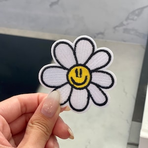 May include: White embroidered iron-on patch of a daisy with a yellow smiley face in the center.