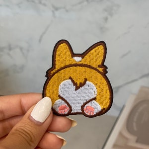 Könnte beinhalten: Ein gestickter Aufnäher mit dem Hinterteil eines Corgis, mit braunem und gelbem Fell, einem weißen Schwanz und rosa Pfoten.