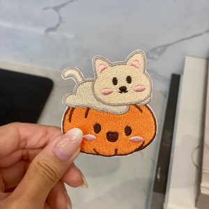 Puede incluir: Parche bordado con un gato color crema acostado sobre una calabaza naranja. El gato tiene acentos rosas en las orejas y las mejillas, y la calabaza tiene una cara sonriente. El parche tiene un borde cosido.
