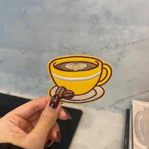 Könnte beinhalten: Ein gelber gestickter Bügelaufkleber mit einer Kaffeetasse mit einem Herzdesign im Schaum und einer Kaffeebohne auf einem Unterteller.
