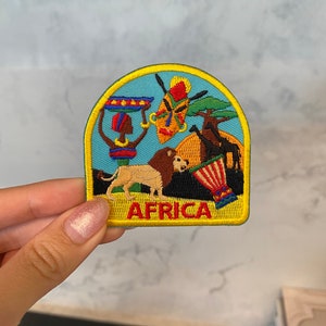 Può includere: Una toppa ricamata gialla, blu e verde con un leone, un tamburo, una maschera e un albero. Il testo "AFRICA" è scritto in giallo su sfondo nero.