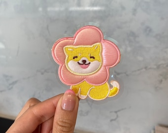 Puede incluir: Un perro amarillo bordado con un parche en forma de flor rosa. El parche está sobre un fondo transparente.