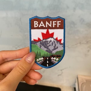 May include: A blue, brown, white, green, and red embroidered patch with the word "BANFF" in white letters on a brown background. The patch features a mountain range with a road and a Canadian maple leaf.