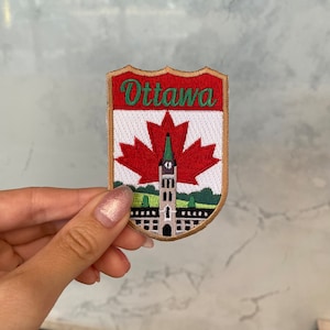 Puede incluir: Un parche bordado de color rojo, blanco y verde con la palabra "Ottawa" en letras verdes. El parche presenta una hoja de arce roja con un contorno blanco y un edificio con una torre de reloj en el centro. El parche se sostiene en una mano.