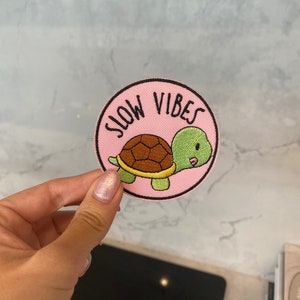 Puede incluir: Un parche bordado de color rosa y blanco con una tortuga de dibujos animados y el texto "Slow Vibes".