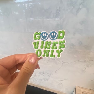 May include: Embroidered patch with the words "GOOD VIBES ONLY" in green, with two blue smiley faces. The patch has a white background and a textured appearance. The patch is a fun accessory.