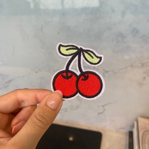 Puede incluir: Un parche bordado de cerezas rojas y verdes con costuras blancas.