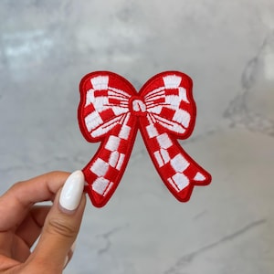 Könnte beinhalten: Ein rot-weiß kariertes Schleifen-Patch mit rotem Rand. Die Schleife ist mit einem klassischen Karomuster bestickt. Der Aufnäher wird von einer Hand mit weißem Nagellack gehalten.