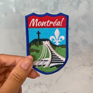 May include: A blue, green, and white embroidered patch with the text "Montréal" in white. The patch features a stylized image of a bridge, a cross, and a fleur-de-lis.