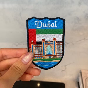 Op de afbeelding: Een geborduurde patch in blauw, wit, groen en rood met het woord "Dubai" in witte letters. De patch toont een gebouw met een grote boog en een blauwe koepel.