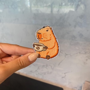 May include: A brown and orange embroidered patch of a capybara holding a coffee cup. The capybara is sitting down and has a brown nose and eyes.