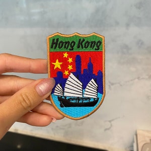 May include: A red, green, and blue embroidered patch with the text "Hong Kong" in black. The patch features a yellow five-pointed star on a red background, a blue skyline, and a white sailboat on a blue sea.