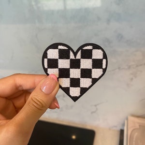 Puede incluir: Un parche en forma de corazón a cuadros en blanco y negro. El parche está hecho de tela y tiene un borde cosido.