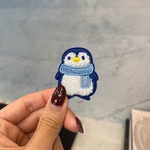 May include: A blue and white penguin with a blue scarf embroidered patch. The penguin is wearing a blue scarf and has a yellow beak and eyes.