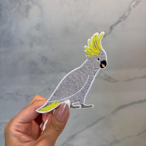 May include: Embroidered patch of a gray cockatoo bird with a yellow crest and tail accents. The bird is depicted in a side profile, with detailed stitching and a white border. The patch is designed for application on clothing or accessories.
