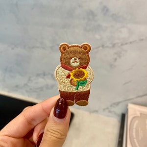 Könnte beinhalten: Gestickter brauner Bären-Patch, der eine gelbe Sonnenblume hält. Der Bär trägt einen cremefarbenen Pullover mit einem roten Schal.