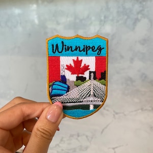 May include: A blue and gold embroidered patch with the word "Winnipeg" in black lettering. The patch features a Canadian flag with a red maple leaf and a depiction of the city's skyline, including the Canadian Museum for Human Rights and the Esplanade Riel.