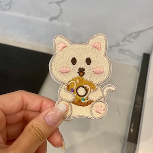 Könnte beinhalten: Ein weiß gestickter Bügelaufkleber mit einem Cartoon-Katzenmotiv, das einen braunen Donut mit bunten Streuseln hält.