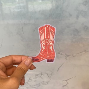 Puede incluir: Un parche bordado rosa de una bota vaquera con costuras blancas.