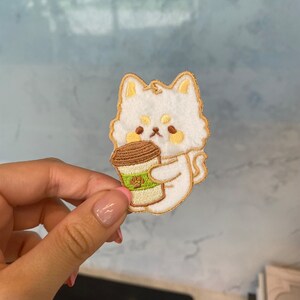 Op de afbeelding: Witte geborduurde patch van een cartoonhond die een bruin en groene koffiebeker vasthoudt. De hond heeft gele oren en een bruine neus.