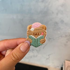 May include: A brown bear wearing a pink hat is reading a green book. The bear is embroidered on a white fabric.