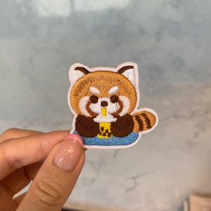 Könnte beinhalten: Gestickter Bügel-Patch mit einem Roten Panda, der einen Boba-Tee hält. Der Rote Panda ist braun und weiß mit einem blauen und weißen Hintergrund.