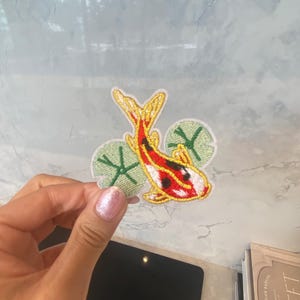 May include: Embroidered iron-on patch featuring a red and white koi fish swimming through green lily pads.