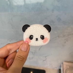Peut inclure: Un écusson en forme de visage de panda blanc avec des oreilles et des yeux noirs et des joues roses. L'écusson est tenu par une main. L'écusson panda est un accessoire mignon pour les vêtements, les sacs ou autres articles.