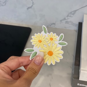 Peut inclure: Écusson brodé représentant un bouquet de marguerites jaunes avec des centres orange et des feuilles vertes. L'écusson est sur fond blanc. L'écusson est tenu par une personne.