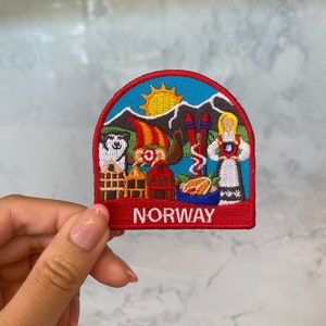 May include: Embroidered patch featuring a Norwegian landscape with mountains, a sun, and traditional figures. The patch has a red border with the word "NORWAY" at the bottom. The design includes a dog, a gnome, and a woman in traditional dress.
