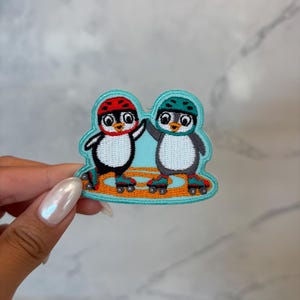 May include: Embroidered patch featuring two cartoon penguins wearing helmets and roller skates. One penguin is black and white with a red helmet, the other is gray and white with a teal helmet. They are on a light blue background.