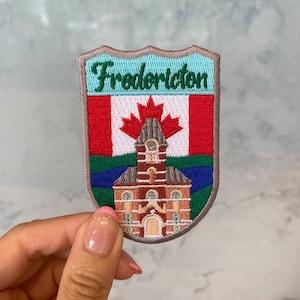 Könnte beinhalten: Gestickter Aufnäher mit einer stilisierten Darstellung eines Gebäudes mit einem Uhrturm, einer kanadischen Flagge und dem Text "Fredericton". Der Aufnäher ist in den Farben Rot, Weiß, Blau, Grün und Braun gehalten.