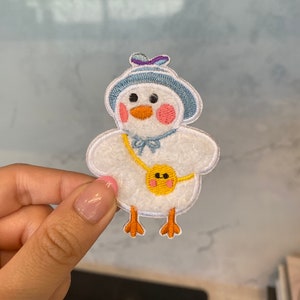 Puede incluir: Parche blanco bordado para planchar de un pollito de dibujos animados que lleva un sombrero azul con un lazo morado, una bolsa amarilla y pies naranjas.