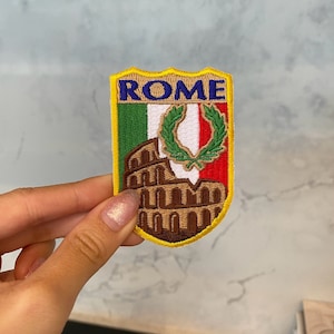 Op de afbeelding: Een geborduurde patch in geel, bruin, groen, wit en rood met het woord "ROME" in blauwe letters. De patch toont een afbeelding van het Colosseum en de Italiaanse vlag.