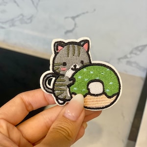 Könnte beinhalten: Ein gestickter Aufnäher mit einer grauen und weißen Katze, die einen grünen und weißen Donut mit weißen Streuseln hält. Der Donut hat einen braunen Zuckerguss.