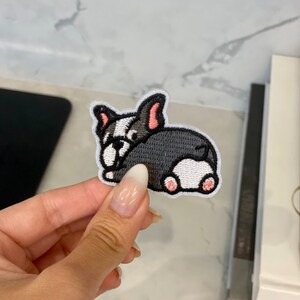 Può includere: Un toppa ricamata da stirare con un bulldog francese di cartone animato, in grigio e bianco con orecchie e zampe rosa.