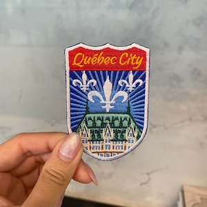 Könnte beinhalten: Ein blauer und roter gestickter Aufnäher mit dem Text "Quebec City" in Gelb. Der Aufnäher zeigt eine weiße Fleur-de-lis und ein stilisiertes Gebäude mit einem grünen Dach.