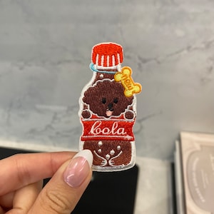 Pode incluir: Etiqueta bordada de um poodle marrom dentro de uma garrafa de cola vermelha e branca com a palavra "boola" no rótulo. Um osso amarelo com a palavra "chew" está no pescoço da garrafa.