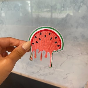 Puede incluir: Un parche de hierro bordado con forma de sandía roja y verde con efecto de goteo. La sandía tiene forma de semicírculo con semillas negras.