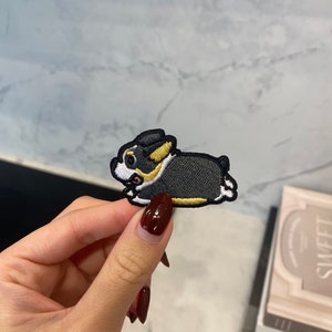 May include: Black and white embroidered patch of a cartoon dog with yellow accents. The dog is running and has a happy expression.