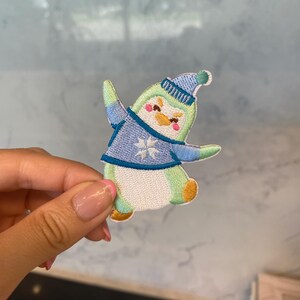 May include: Embroidered patch featuring a cartoon penguin in a winter outfit. The penguin is wearing a blue hat and sweater with a snowflake design. The penguin has a white belly, green body, and orange feet and beak. The patch is held by a hand.