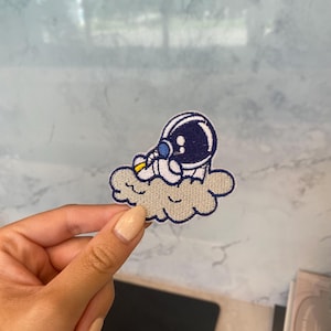 May include: Embroidered patch featuring an astronaut in a navy blue helmet and white suit, seated on a gray cloud. The patch has yellow and blue accents. The design is a whimsical depiction of space travel.