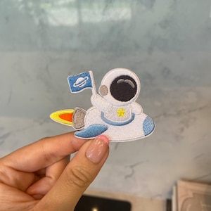 May include: A white astronaut with a black face and a yellow star on its chest is flying in a blue and white spaceship with a yellow flame. The astronaut is holding a blue flag with a white planet on it.