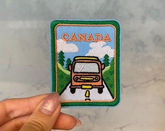 Puede incluir: Un parche bordado azul y verde con un autobús de dibujos animados amarillo y naranja conduciendo por un camino blanco con la palabra "CANADA" en letras naranjas.