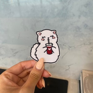 Può includere: Toppa ricamata bianca da stirare con un gatto dei cartoni animati con guance rosa e occhi chiusi che tiene una tazza rossa con una cannuccia. Il gatto ha un contorno nero.