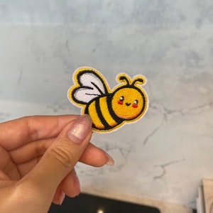 Op de afbeelding: Een geborduurde bij-patch in geel en zwart met een witte vleugel en een glimlachend gezicht. De bij houdt een klein rood hart vast.