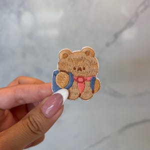 May include: Embroidered patch featuring a brown teddy bear holding a blue and white object, with a pink bow. The patch has a stitched design and is likely for clothing or accessories.