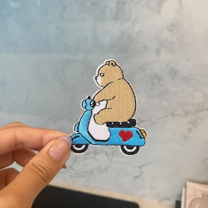 May include: A brown bear riding a blue scooter with a red heart on the side. The scooter has white wheels and a white seat.