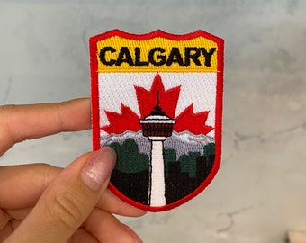 Puede incluir: Parche bordado en rojo, blanco y amarillo con la palabra "CALGARY" en letras negras. El parche presenta una hoja de arce roja con una torre blanca en el centro y un paisaje urbano verde y negro debajo.