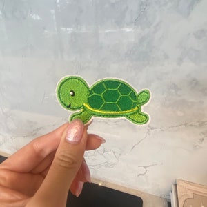 Può includere: Toppa di tartaruga verde ricamata con contorno giallo. La tartaruga ha un viso sorridente e un guscio con un motivo verde e giallo.